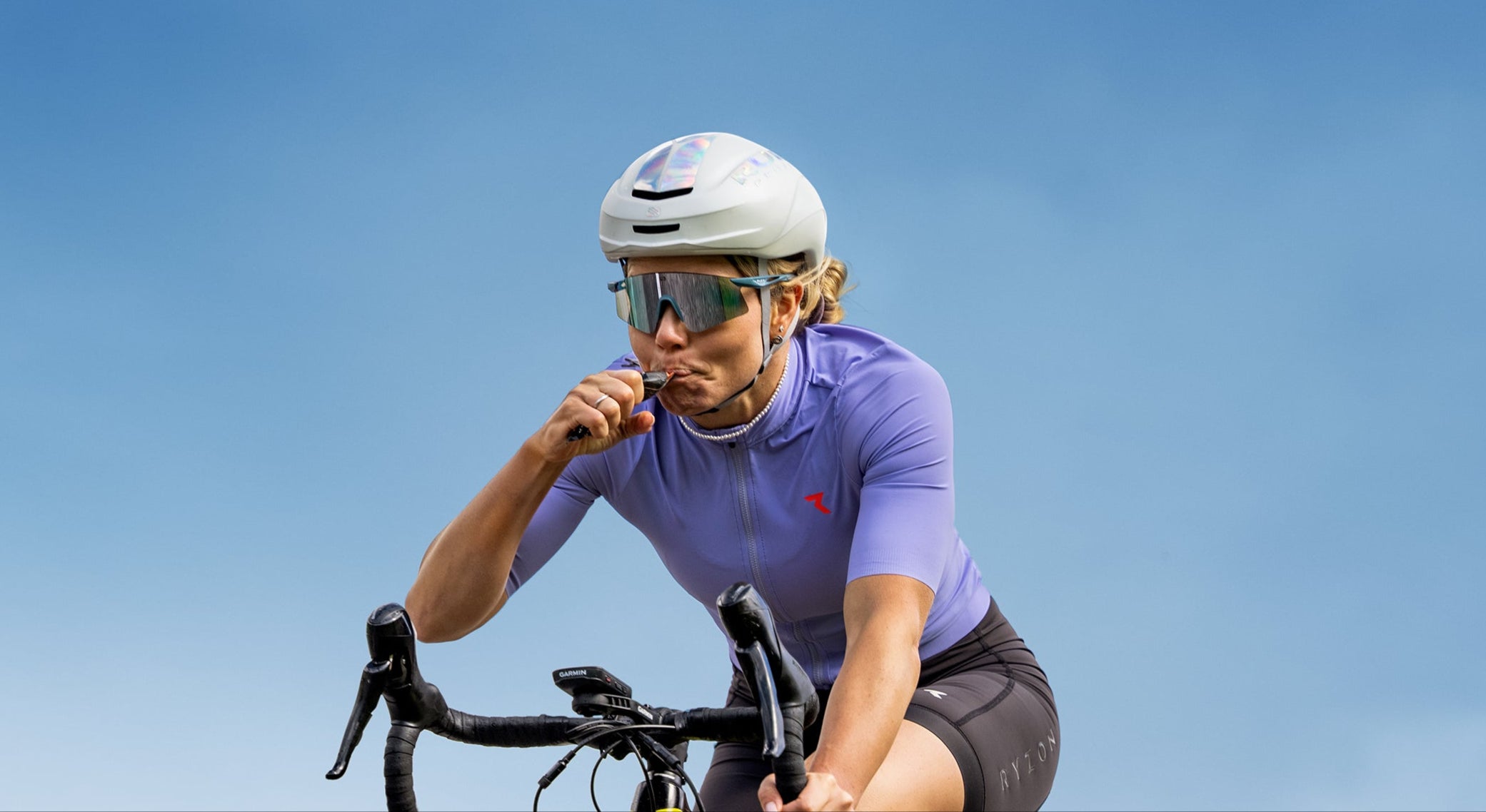 Cyclist riding a road bike refueling with a Powerbar energy gel outdoors under a blue sky during training.